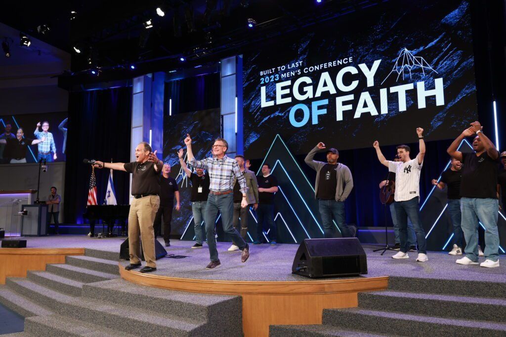 People on a stage with raised hands, under a “LEGACY OF FAITH” sign at a church event