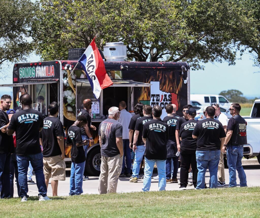 Group of people in black shirts gathered outdoors near a truck and flag at a grassy park.
