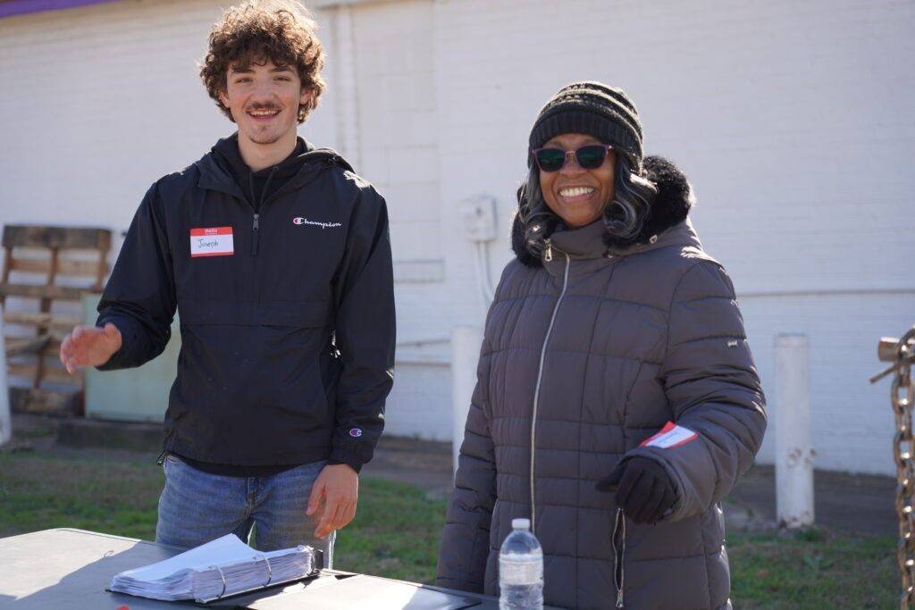 Two people standing at an outdoor table, smiling in winter jackets beside a building
