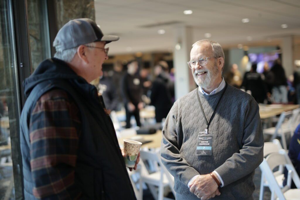 Two men chatting in a bright indoor lobby, one holding a cup and wearing a lanyard.
