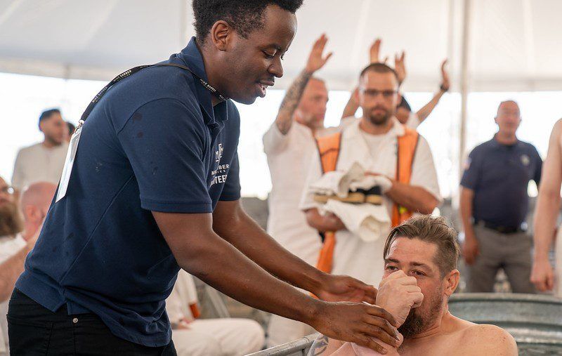 Man adjusting a seated man’s face inside a bright tent while others watch in the background