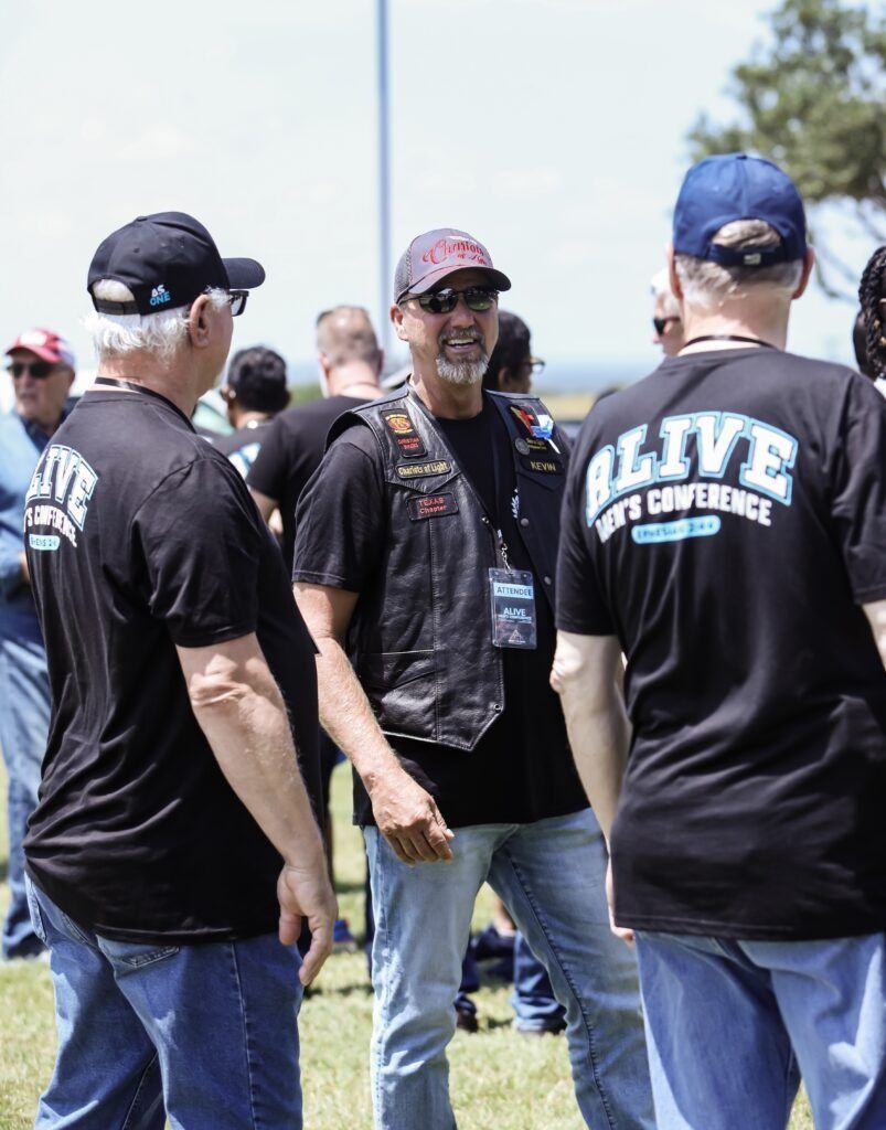People in black event T-shirts and caps standing outdoors in a sunny grassy area.