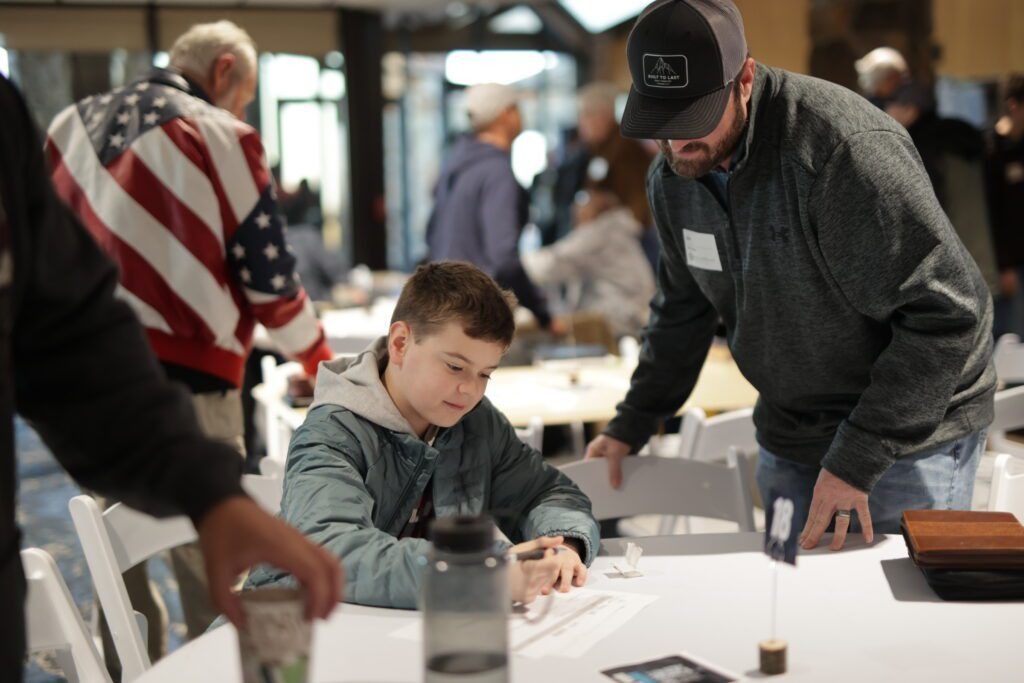 Two people leaning over a table helping a child write at a bright indoor event booth
