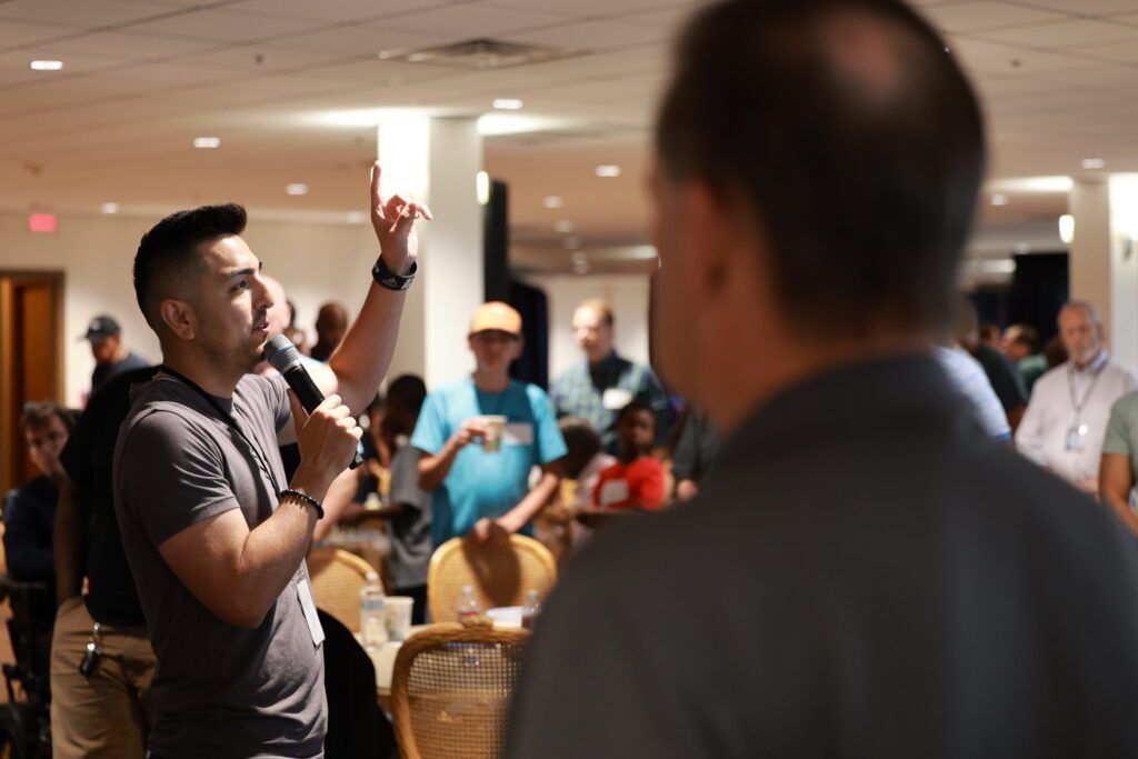 Man speaking into a microphone at a conference, with attendees seated in the background