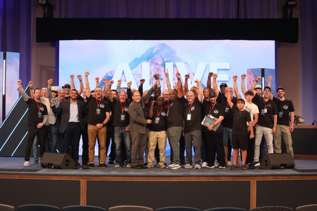 Group of people on a stage raising their arms in celebration in front of a large screen and purple curtains