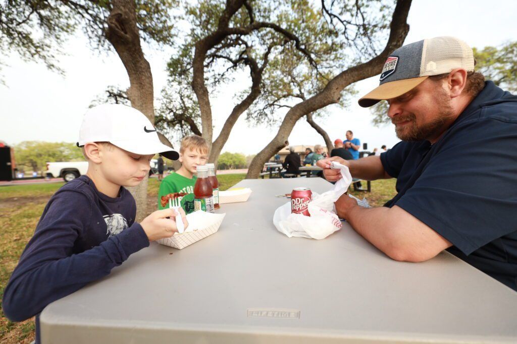 Two people sit at a picnic table outdoors, eating from takeout containers under trees.