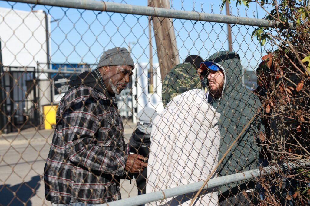 People standing by a chain-link fence, carrying a large white bag outdoors.