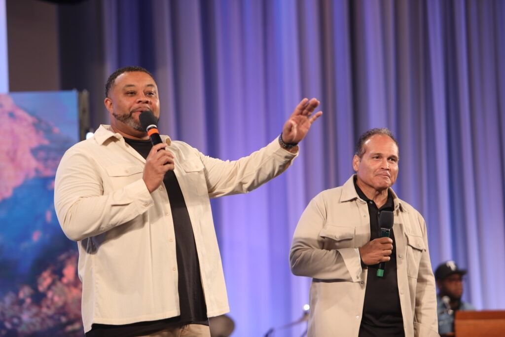 Two men speaking onstage with microphones before blue curtains