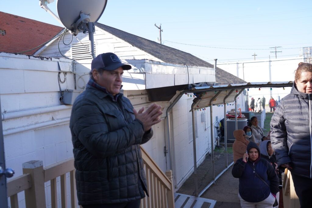 People standing on a snowy balcony beside white buildings, one man clapping near a satellite dish.
