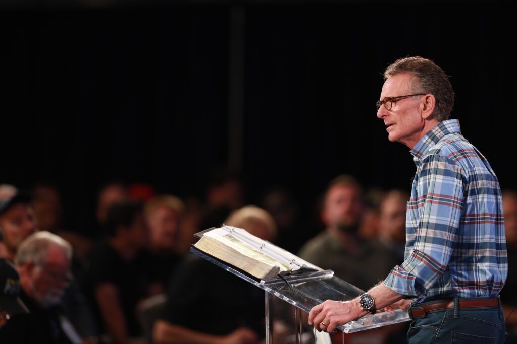 Man in plaid shirt speaking at a podium, with an open book, in front of a seated audience