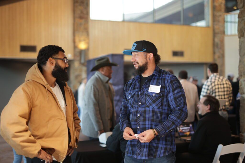 Two men talking indoors at a casual gathering, with people seated in the background.