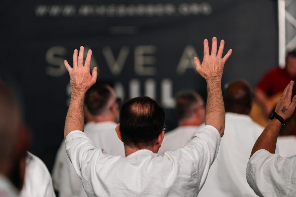 People with raised hands facing a stage with a blurred “SAVE A …” banner in the background