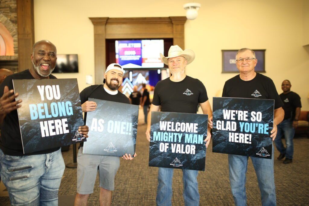 Four men indoors hold signs reading “YOU BELONG HERE,” “WE ARE ONE,” and “WELCOME MIGHTY MAN OF VALOR.”