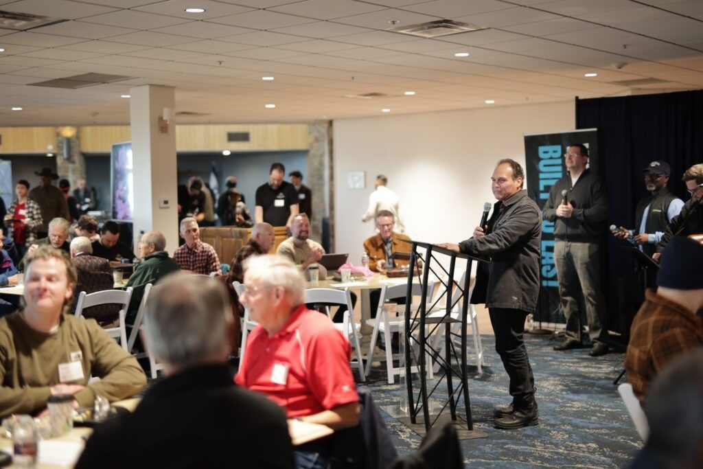 People gathered in a hotel ballroom, with a speaker standing near tall tables and a crowd seated around them.
