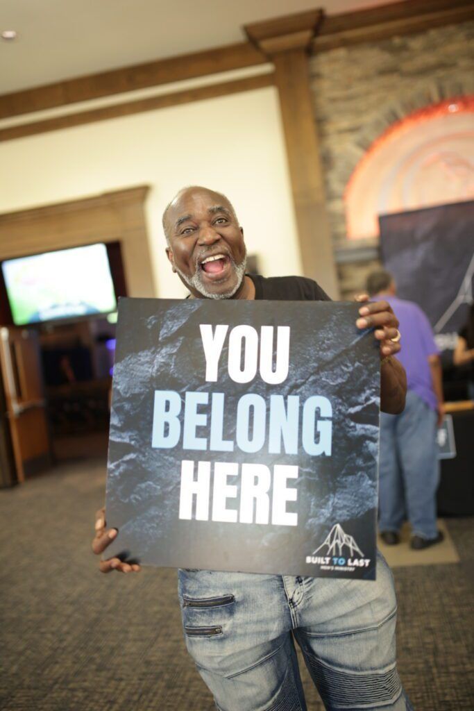 Man holding a sign reading “YOU BELONG HERE” at an indoor event, smiling at the camera.