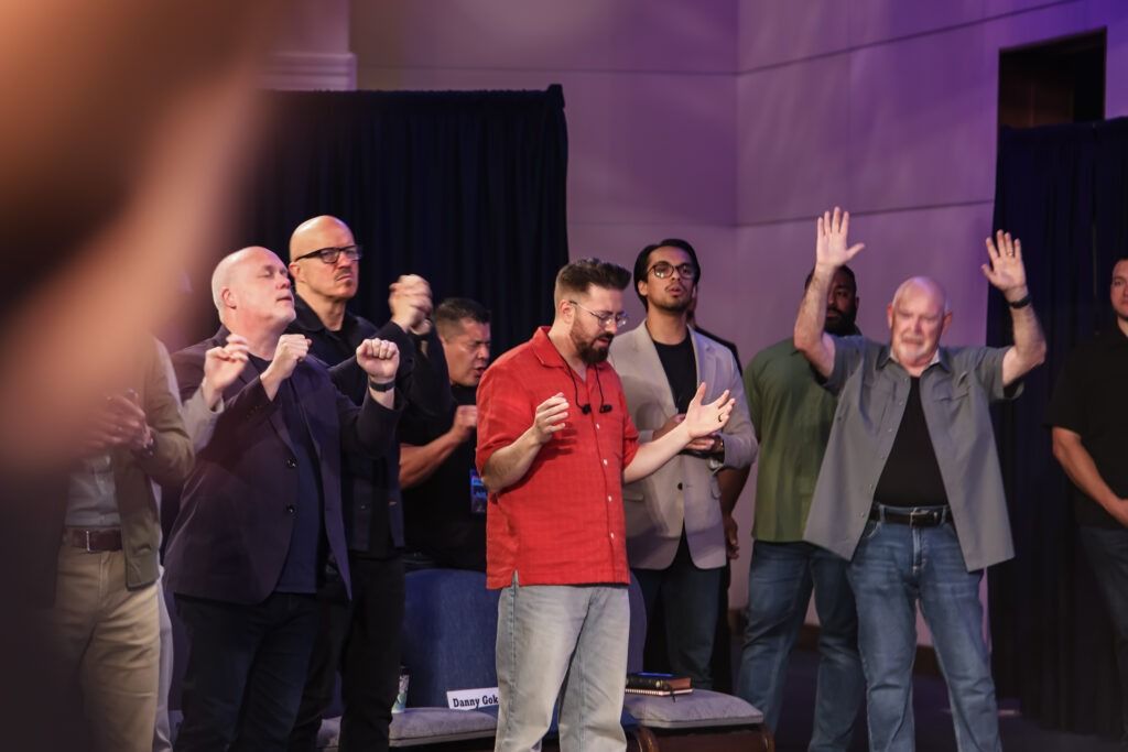People on a stage raising their hands, one man in a red shirt holding a microphone, under purple lighting
