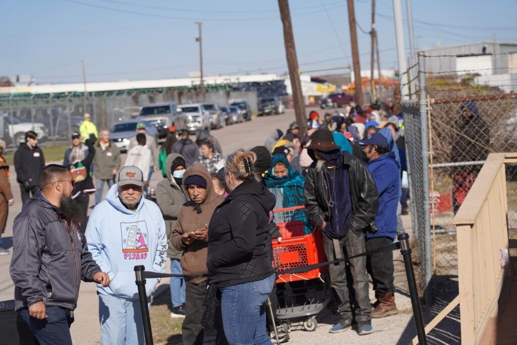 Group of people gathered outdoors near a chain-link fence, many wearing winter coats and hats, waiting or talking.