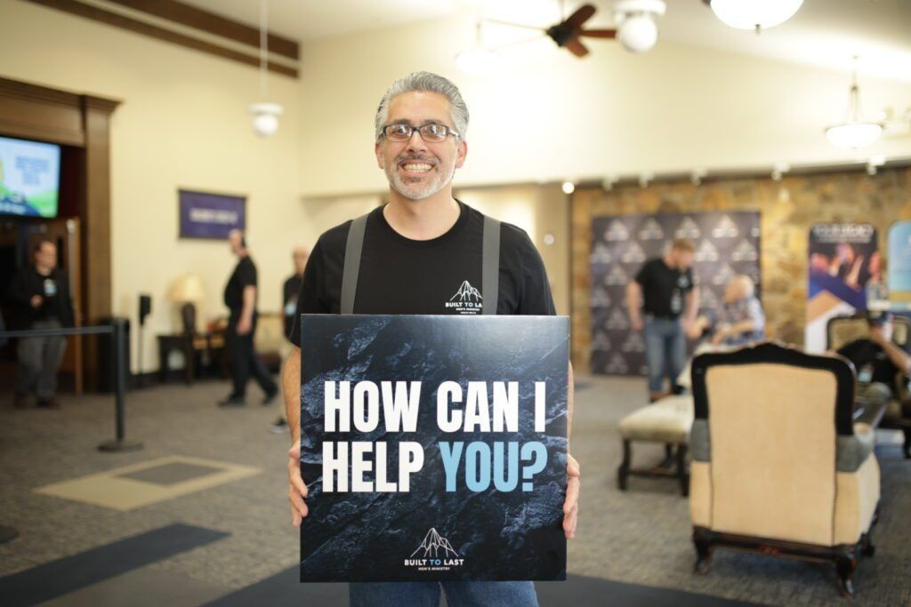 Man holding a sign that says “HOW CAN I HELP YOU?” in a bright indoor lobby