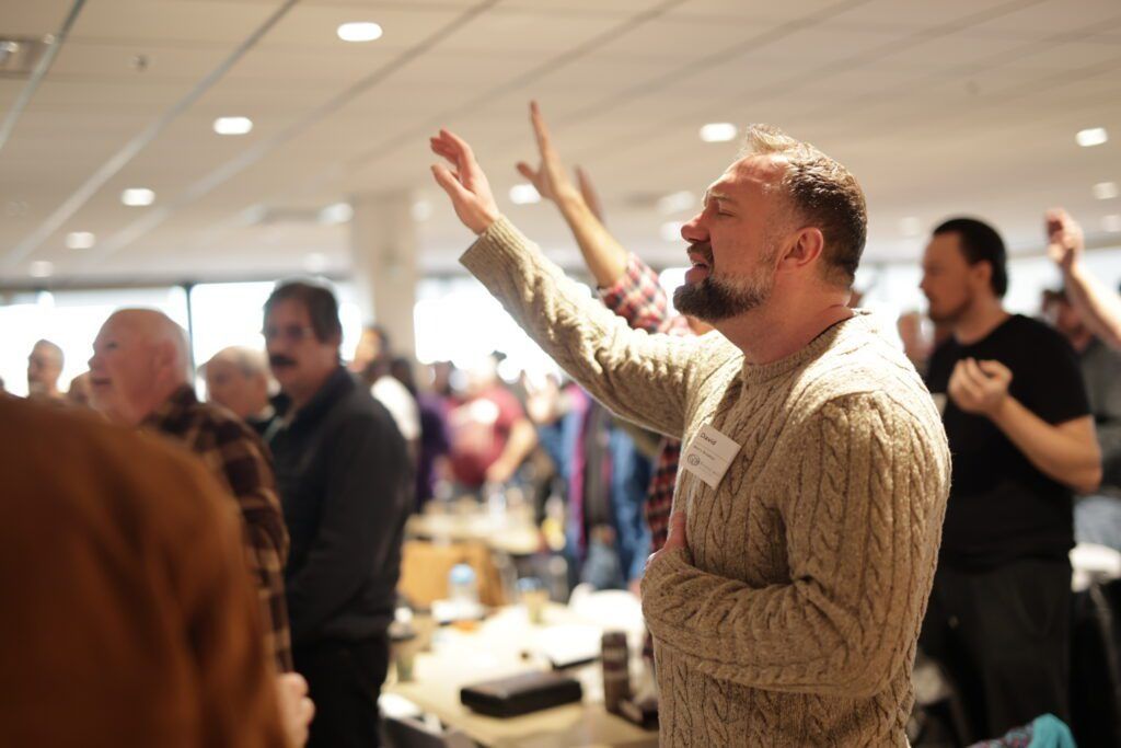 Man in a beige sweater raising both hands in a busy indoor event space