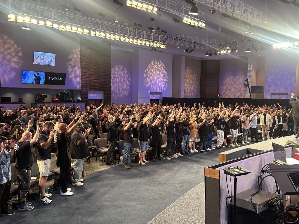 Large crowd standing with raised hands in a purple-lit conference hall, facing a stage and screens.