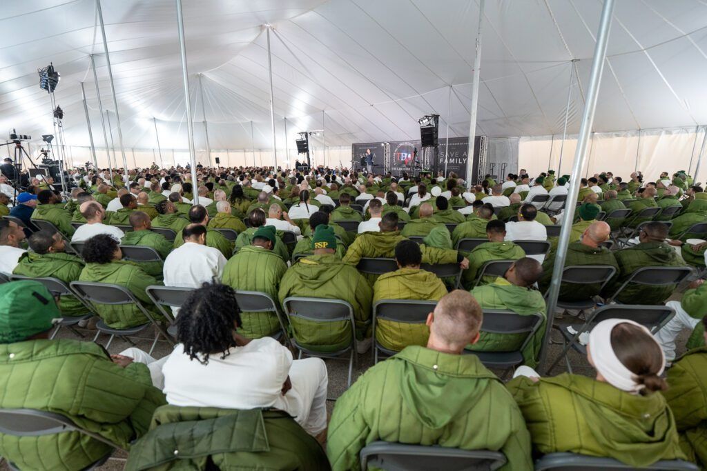 Large indoor gathering in a white tent, with rows of people seated facing a stage.
