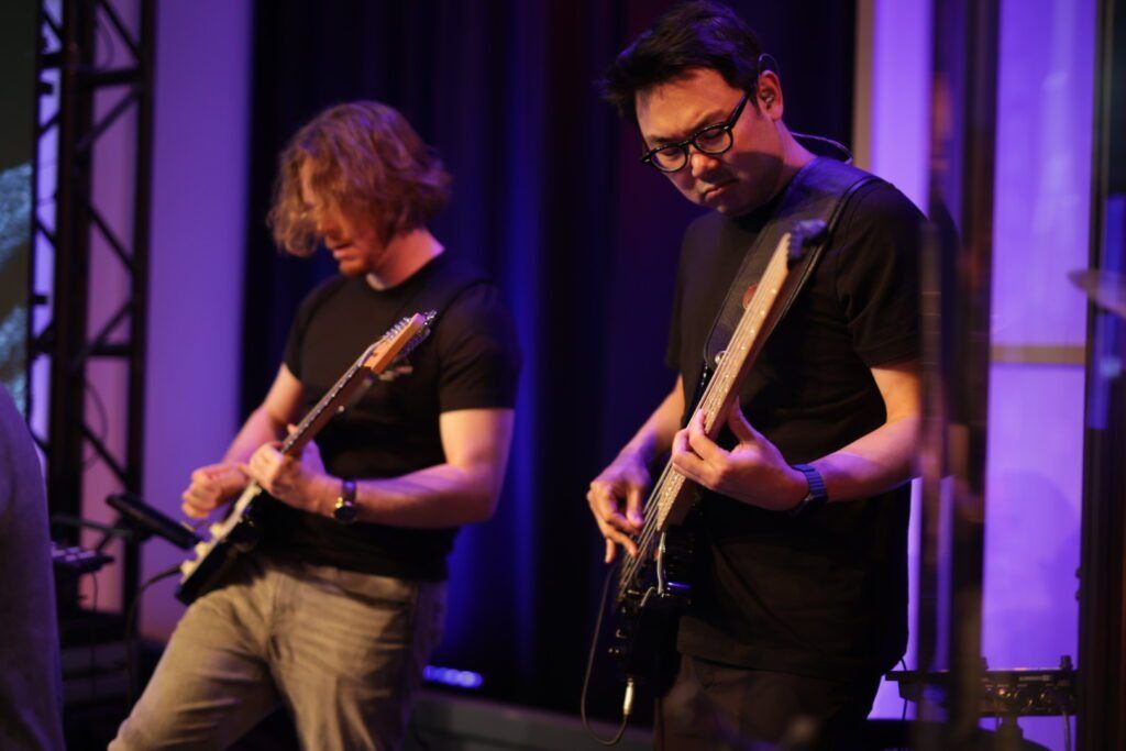 Two musicians playing electric guitars onstage under purple lighting