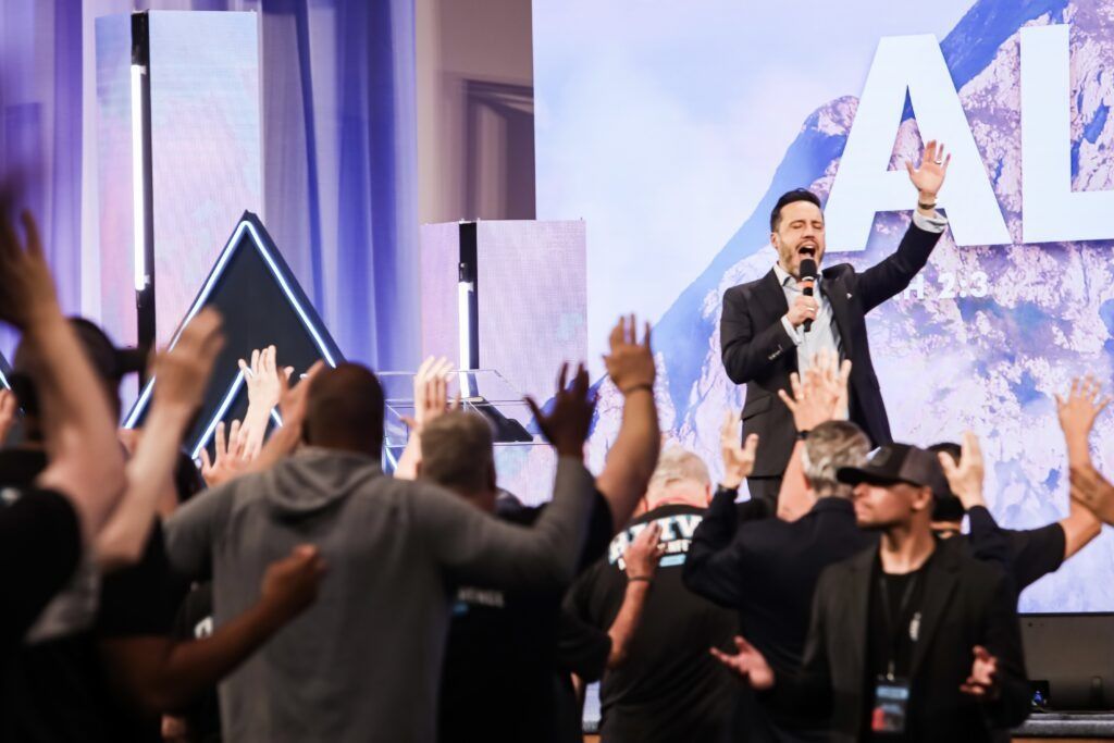 Speaker on stage leading a worship crowd with raised hands under bright lights