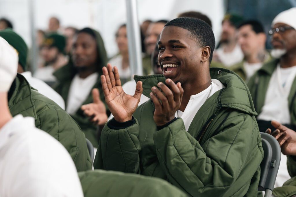 Smiling man in a green jacket clapping at an indoor event