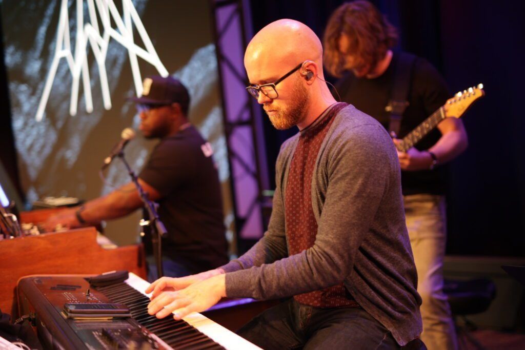 Keyboard player performing on stage with a guitarist in the background under purple lighting