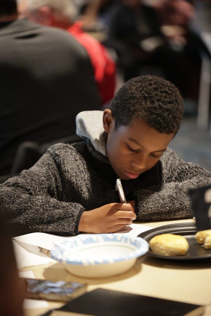 Boy writing at a table with a plate of food and a white bowl in front of him