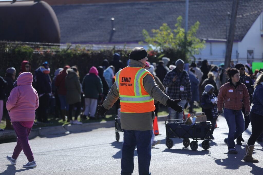 Crowd gathered outdoors near a building, with a safety worker in a reflective vest directing people.