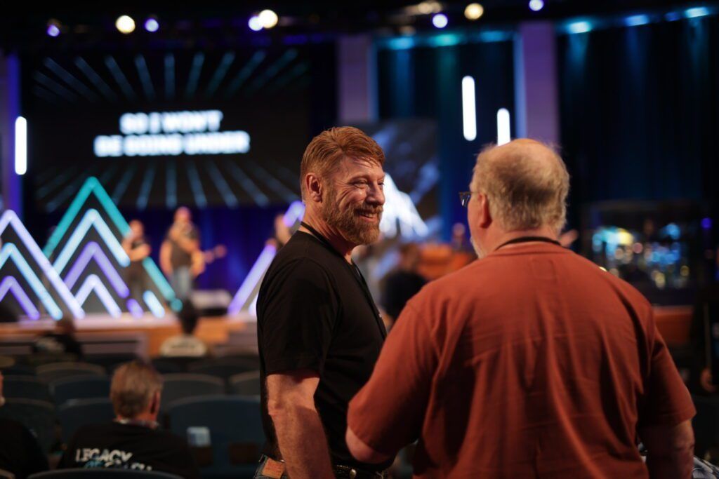 Two men talking in a dim event venue with a lit stage and zigzag light decorations in the background