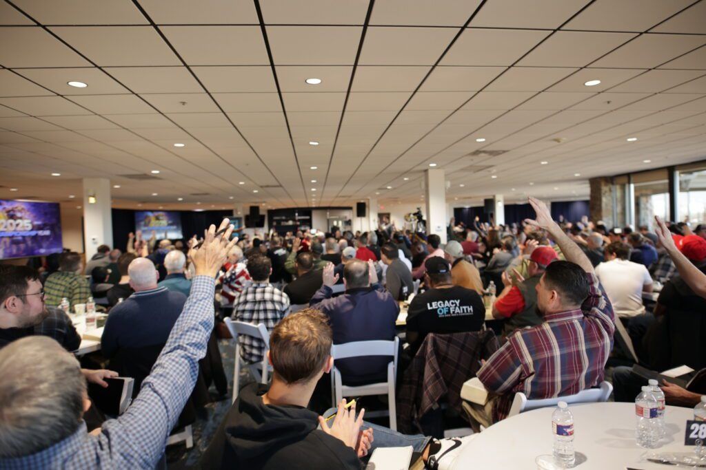 Crowded conference room with seated attendees raising hands during a presentation, viewed from the back