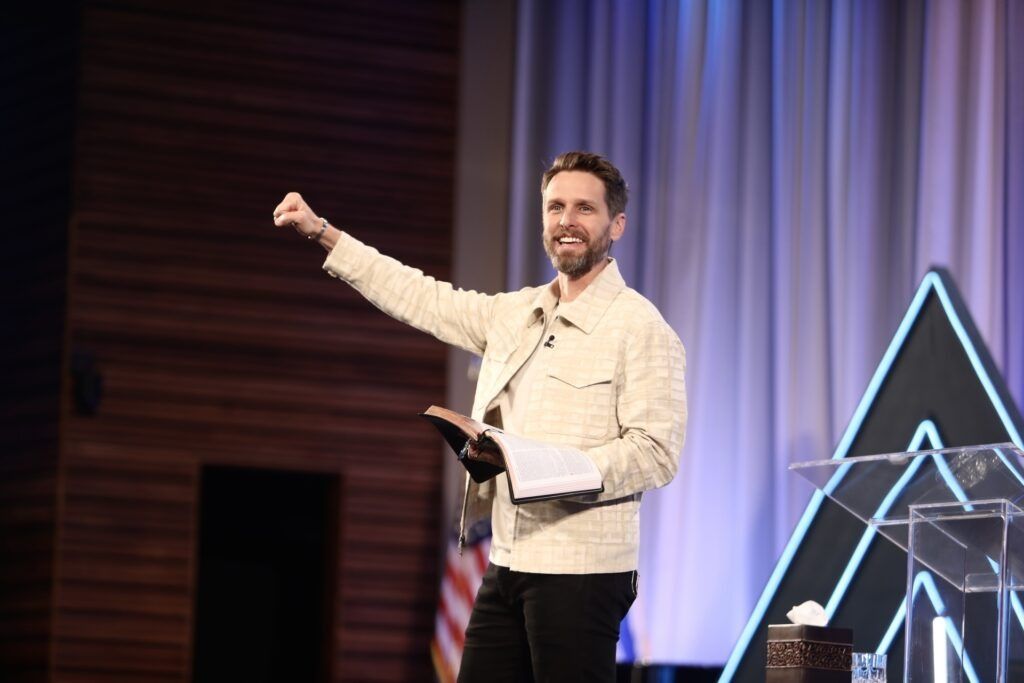 Speaker on stage raising a fist, holding notes, with curtains and glowing geometric backdrop.