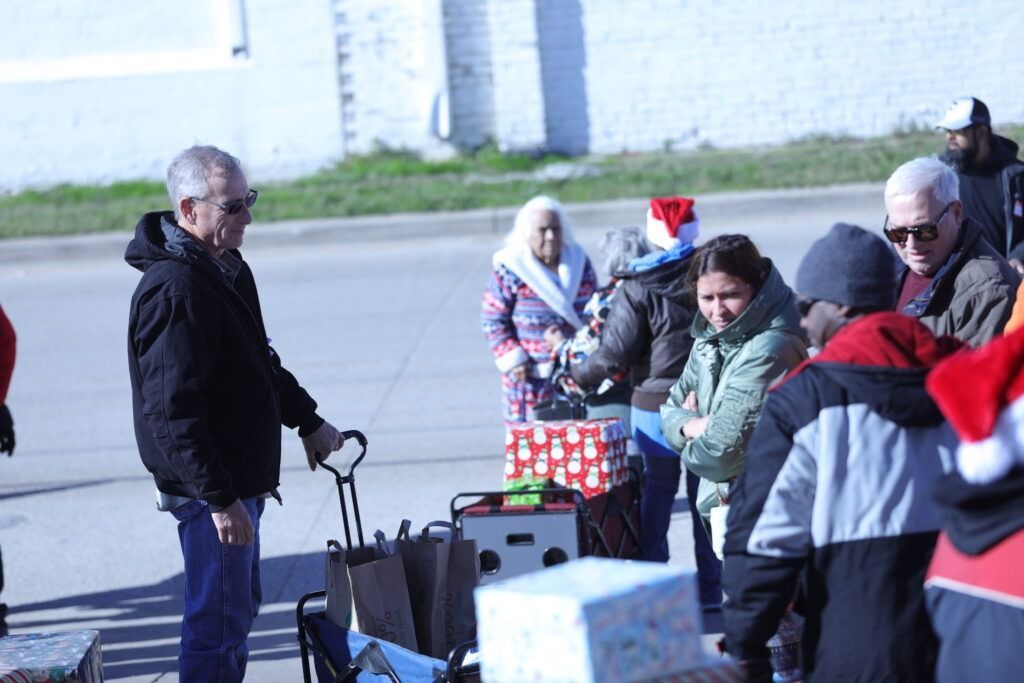 People gathered outdoors with bags and boxes, standing near a man pulling luggage on a cart.