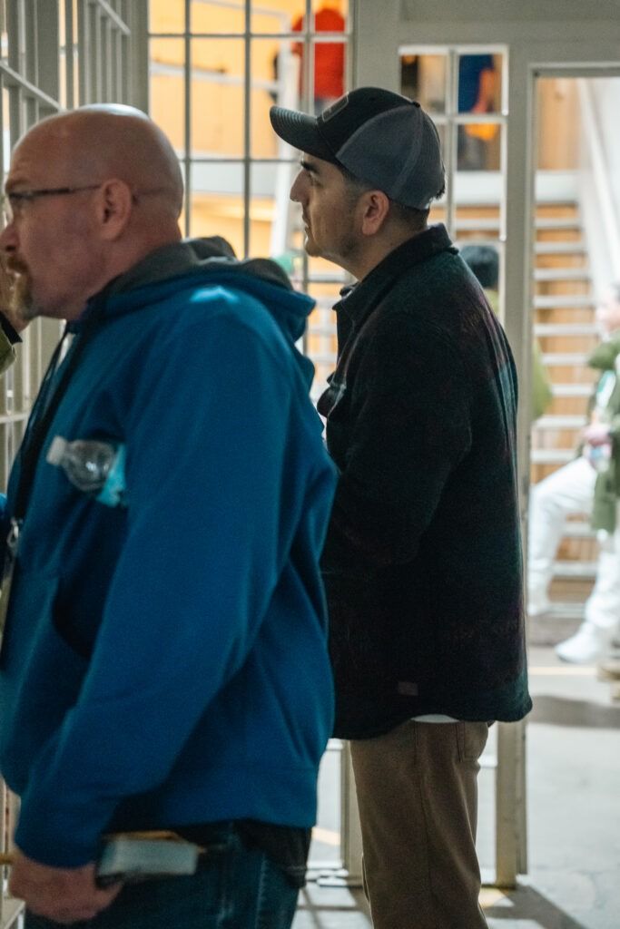 Two men standing indoors near a doorway, one in a blue hoodie and one in a black jacket and cap.