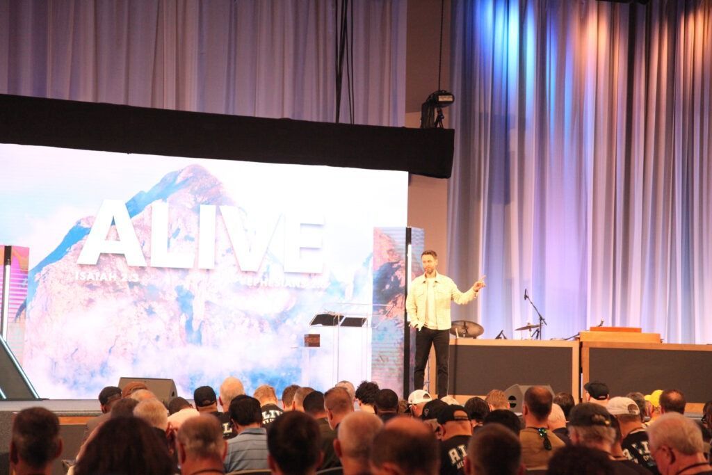 Speaker on stage addressing a crowd in a large auditorium with a bright presentation screen behind him