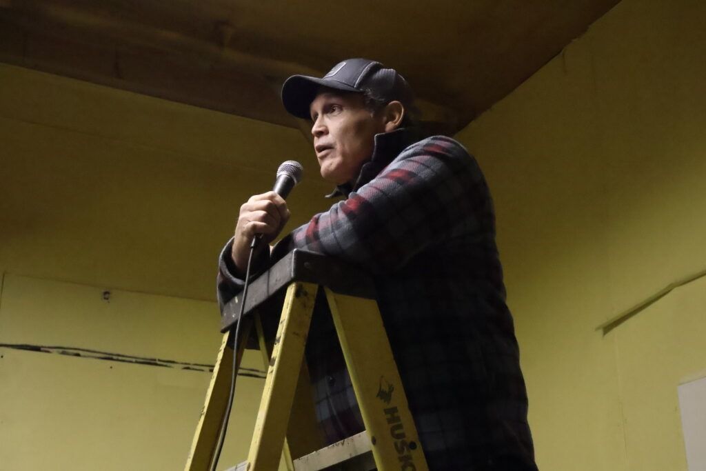 Person speaking into a microphone while standing on a yellow ladder in a room with yellow walls.