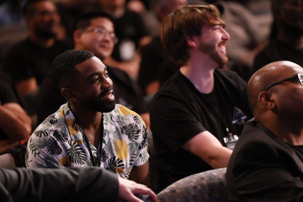 Audience members seated and watching attentively at an event, with one man in a patterned shirt in the foreground