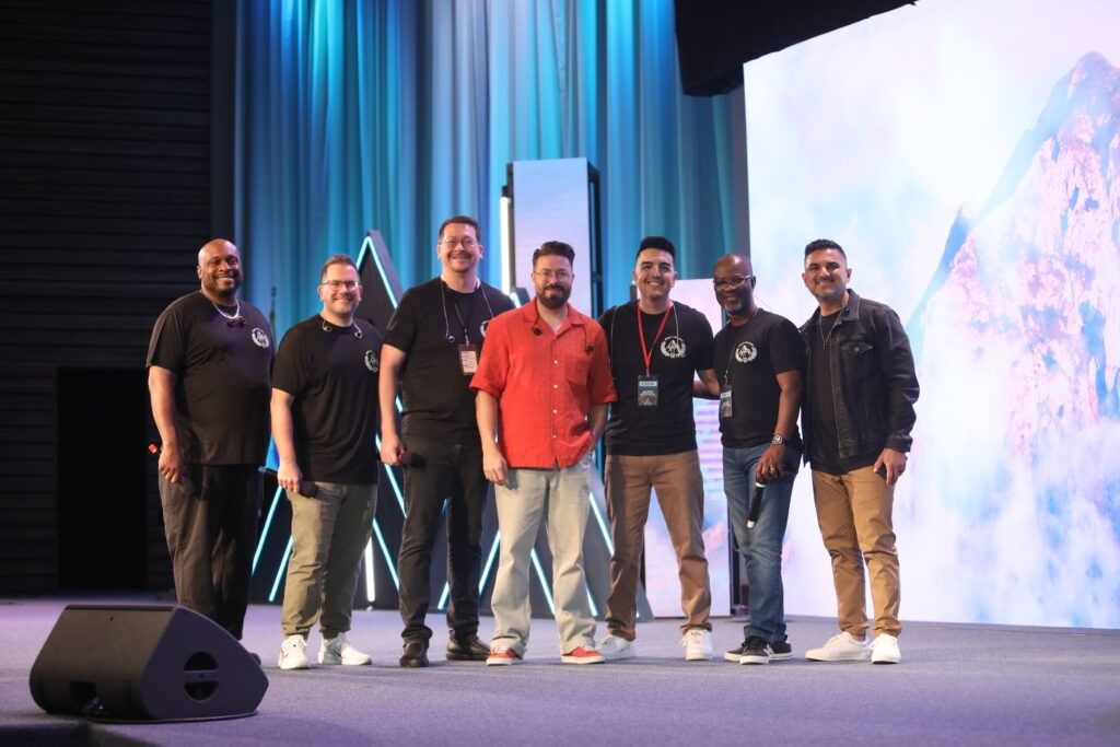 Seven people standing on a stage with blue curtains and a large screen backdrop, one in a red shirt.