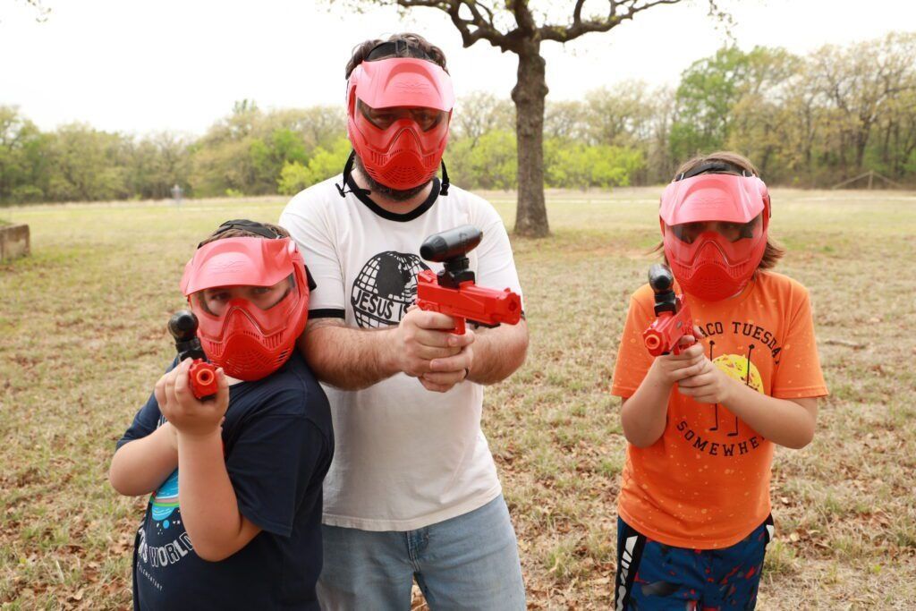 Three people wearing red face shields aim red toy guns outdoors in a grassy field.