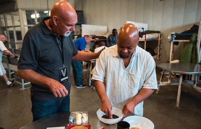 Two people prepare food together at a kitchen counter, one mixing a bowl while the other watches.