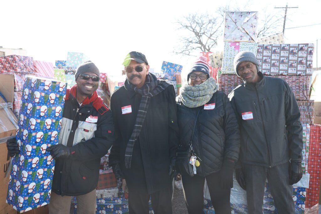 Four people in winter coats standing outdoors beside stacks of wrapped boxes and supplies