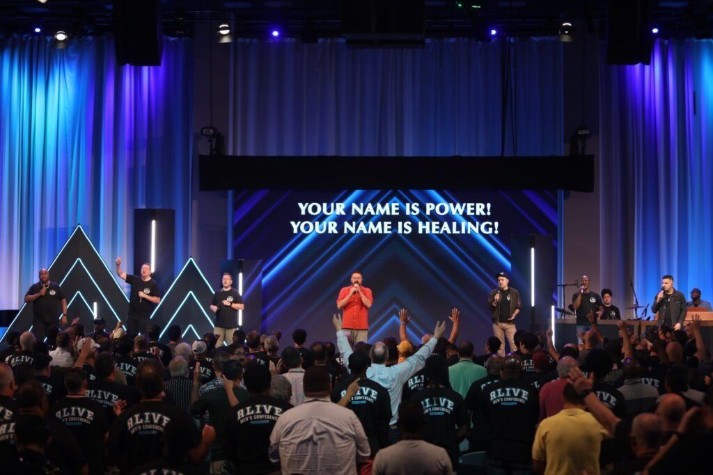 Crowded church stage with blue lighting and screen reading “Your name is power! Your name is healing!”