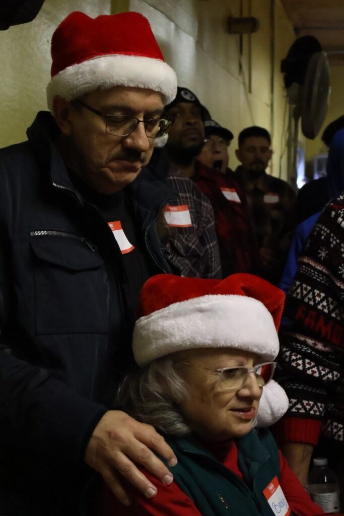 Two people in Santa hats pose indoors, one standing behind the seated person with a hand on their shoulder.