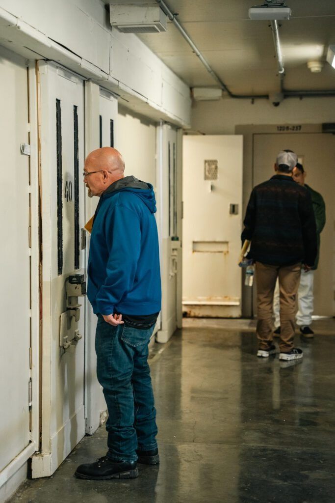 Two men standing in a hallway, one in a blue hoodie near a wall and one farther away in a dark jacket.