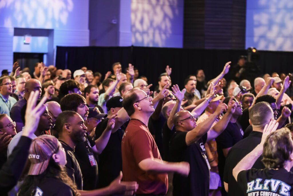 Crowd of people at a conference raising hands in a large indoor venue, lit with purple and blue stage lights