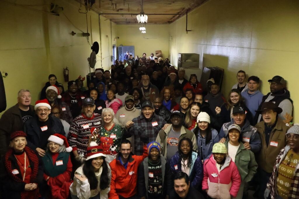 Large group posing in a narrow indoor hallway, many wearing festive holiday sweaters and Santa hats.