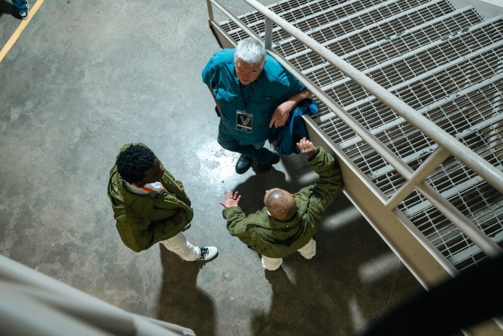 Three people standing in an indoor stairwell beside a metal railing, viewed from above