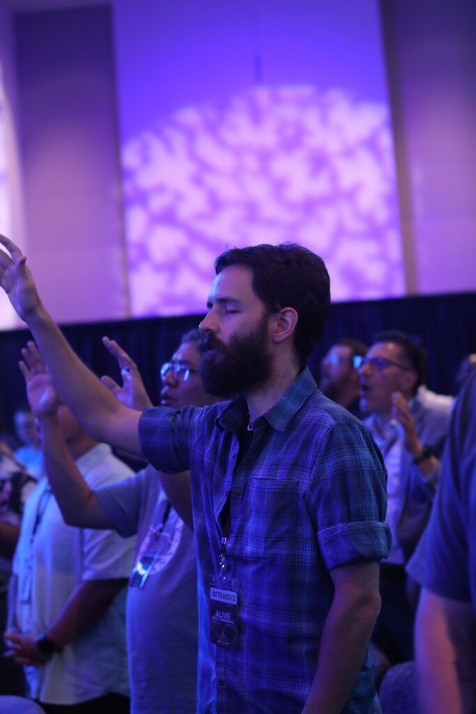 Man with beard in blue plaid shirt raising his hand at a purple-lit indoor event